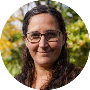 Sarah Lambertson smiling against a nature background. She has brown shoulder-length hair and glasses.