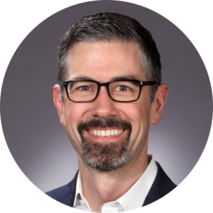 Watt Hamlett, smiling against a gray background. He has dark hair, a beard, and glasses, and is wearing a white shirt and dark jacket.